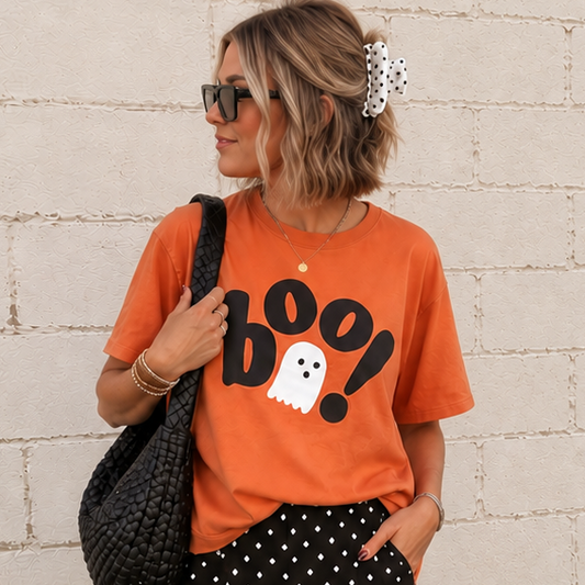 A woman stands against a white painted brick wall wearing an orange graphic T-shirt with a black 'BOO!' and ghost print, black wide-leg pants with white polka dots, and a black woven shoulder bag.