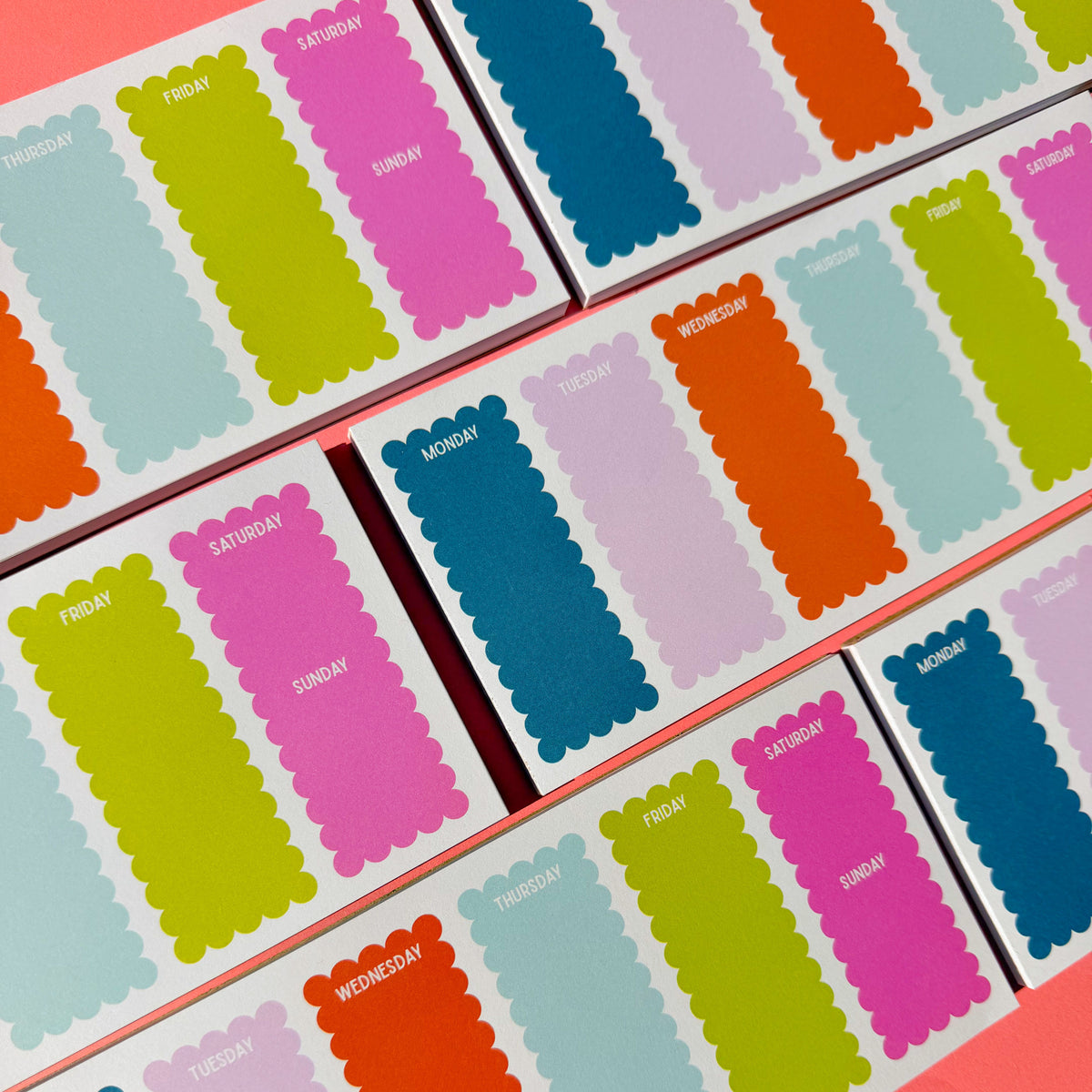 Several small boxes of weekly planner notepads arranged diagonally. They use a scalloped-edge daily labels in pastel and bright colors including blue, green, pink, orange, purple, and light blue, with day names printed on the front.