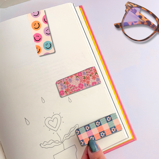 A close-up of a hand holding an open notebook with colorful patterned washi tape strips placed on the pages. A pair of tortoiseshell eyeglasses rests on the right side of the frame.